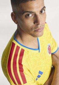Jeune homme les bras croisés porte un maillot de sport jaune avec des bandes rouges et un emblème circulaire sur la poitrine, regardant légèrement vers le haut et vers l'avant.