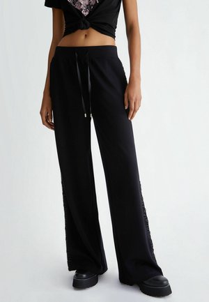 Kvinde iført sorte bukser med vide ben og snøre i taljen samt platformsko, stående mod en ensfarvet lys baggrund.