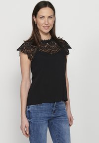 Zwarte blouse met verfijnde kanten kapmouwen, met een hoge halslijn en een vloeiende pasvorm. Draagt met blauwe spijkerbroeken, wat zorgt voor een casual uitstraling.
