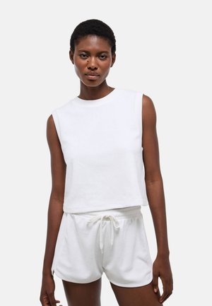 Femme portant un haut blanc sans manches et un short blanc à cordon, debout devant un fond clair uni.