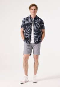 Korte mouwen marineblauw shirt met een wit loofpatroon, gecombineerd met grijze shorts en witte sneakers. Voorzien van een knoopsluiting aan de voorkant en twee borstzakken.