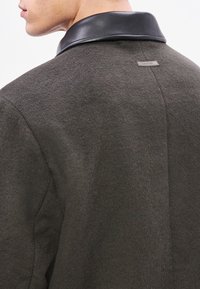 Σκούρο πράσινο μάλλινο παλτό με μαύρο δερμάτινο γιακά. Διαθέτει μια μικρή ετικέτα στο πίσω μέρος. Η υφή φαί νεται μαλακή και ελαφρώς χνουδωτή.