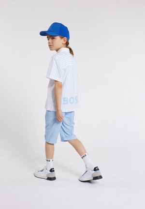 Maglietta bianca a maniche corte con righe blu, pantaloni blu, sneakers bianche con accenti grigi e un cappellino blu. La vista dal retro mostra il logo "BOSS".