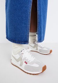 Baskets gris et blanches avec une tige en daim texturé et maille, présentant des accents gris clair, des lacets blancs et une semelle en caoutchouc avec des détails bruns.