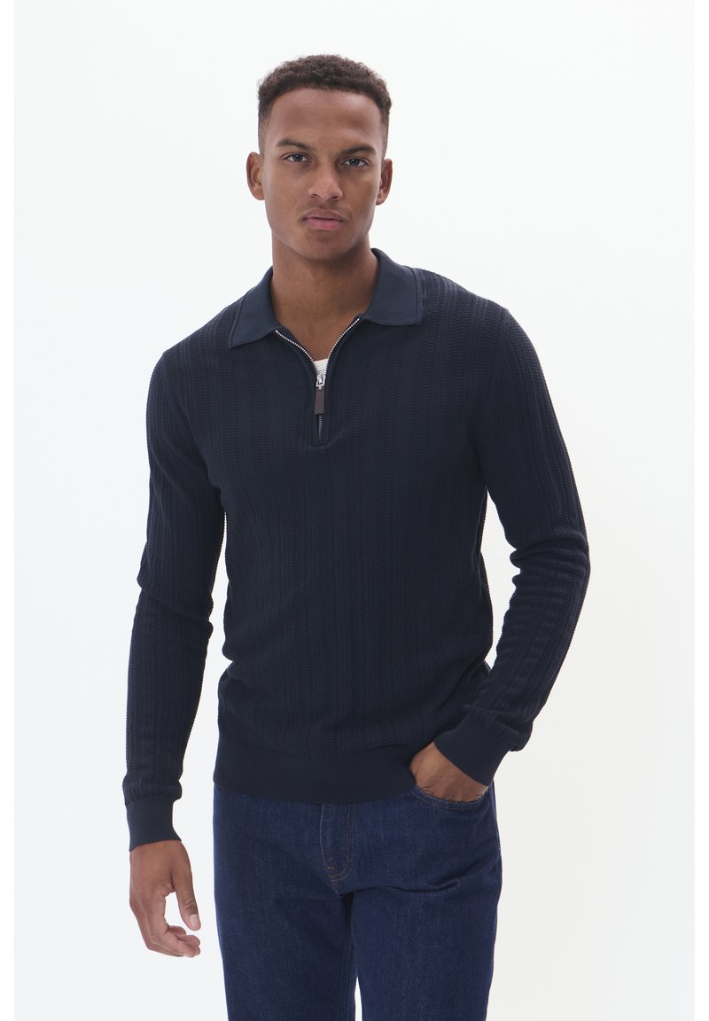 Marinblå stickad tröja med dragkedja i kragen, med ribbstruktur och långa ärmar. Kombinerad med mörka jeans, vilket skapar en åtsittande silhuett.