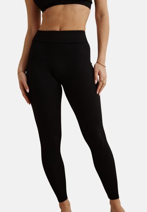 Femme portant un legging noir taille haute et un haut court noir assorti, montrant le ventre et des bijoux dorés aux mains.