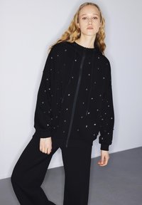 Giovane donna con lunghi capelli biondi ondulati che indossa una felpa nera con zip e piccoli punti metallici, abbinata a pantaloni neri a gamba larga.