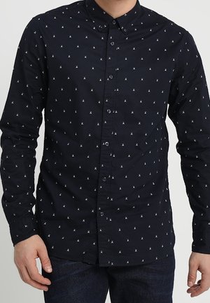 Hombre vistiendo una camisa negra ajustada con botones, con pequeños patrones abstractos blancos y mangas largas.