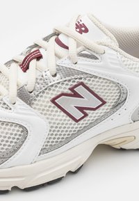 Baskets de sport blanches en mesh avec des accents gris, arborant un logo "N" bordeaux, des lacets crème, des surfaces texturées et une semelle en caoutchouc.