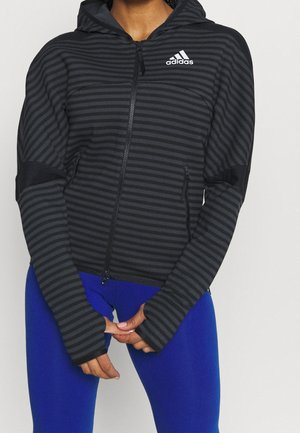 Schwarz-grau gestreifter adidas Zip-up-Hoodie mit Taschen, getragen von einer Person in blauen Leggings, die Hände vor dem Körper verschränkt.