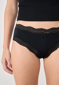 Mutande da donna nere con pizzo in vita, dotate di un delicato fiocco decorativo. Tessuto morbido con una forma classica a slip.