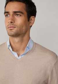 Svetlo béžový sveter s výstrihom do V nosený cez svetlomodrú košelu s golierom. Svetre má hladkú textúru a jemné švy.