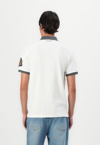 Valkoinen polo-paita, jossa on harmaa kaulus, lyhyet hihat ja logo-patch vasemmassa hihassa. Yhdistetty vaaleansiinisiin farkkuihin.