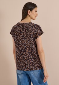 Femme portant un haut à manches courtes à imprimé léopard marron et noir et un jean bleu, vue de dos sur un fond beige uni.