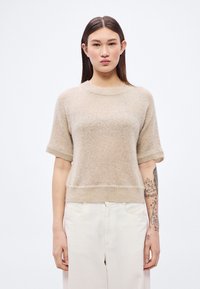 Šviesiai rudas trumpomis rankovėmis megztinis iš minkštos, tekstūruotos medžiagos, su apvalia iškirpte ir raštuotu apačios kraštu. Derinamas su šviesių spalvų kelnėmis.