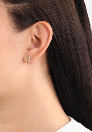 Primo piano di un orecchio femminile con capelli castano scuro che indossa un piccolo orecchino d'argento curvo con tre pietre a forma di stella.
