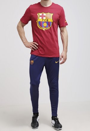 Rotes Baumwoll-T-Shirt mit dem Logo des FC Barcelona, kombiniert mit navyblauen Polyesterhosen, die einen gelben Nike-Swoosh aufweisen. Beide Teile sind in sportlicher Passform.