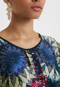 Blusa colorata con grandi motivi floreali blu su un tessuto testurizzato, con scollo ampio e rifiniture verdi, abbinata a orecchini geometrici dorati.
