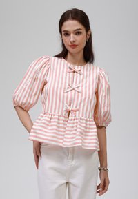 Peplum-Bluse mit rosa-weißen vertikalen Streifen, verfügt über Puffärmel, drei Schleifenakzente und einen runden Ausschnitt. Kombiniert mit weißen Hosen.