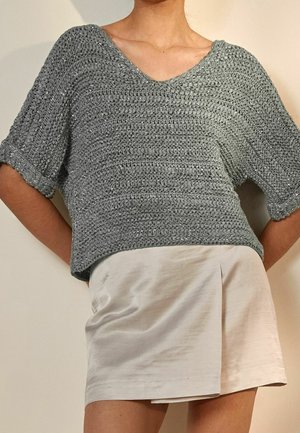 Personne portant un pull gris à col en V en maille lâche et une jupe courte de couleur claire, debout devant un fond uni.