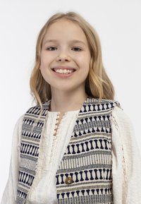 Tkanicowa kamizelka w geometryczne wzory w granatowym i kremowym kolorze, z zapięciem na guziki, noszona na teksturowanej białej bluzce z detalami w postaci guzików.