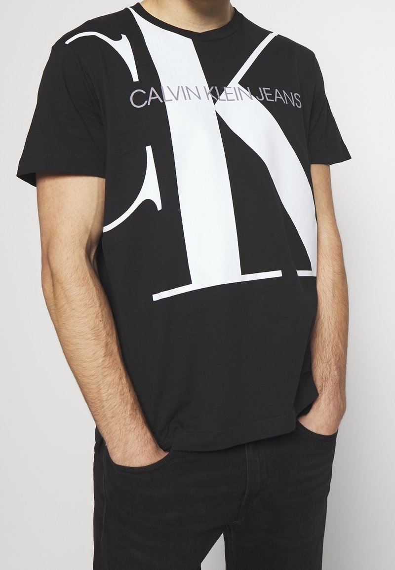 Mies, jolla on päällään musta Calvin Klein -t-paita, jossa on suuri valkoinen "CK"-logo ja "Calvin Klein Jeans" -teksti, kädet taskuissa, tasaisen taustan edessä.