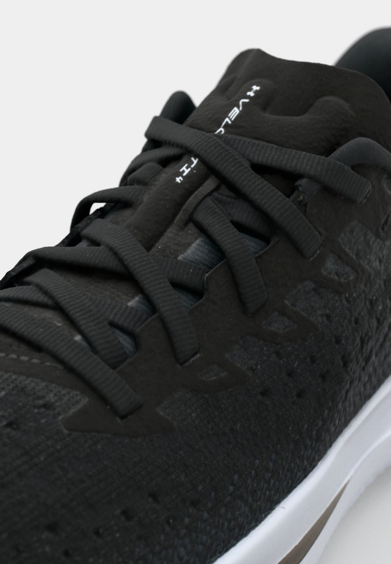 Schwarzer Sportschuh mit strukturiertem Obermaterial, flachen Schnürsenkeln und einer weißen Zwischensohle mit hellbraunen Akzenten. Besonderheiten sind die Belüftungsmuster.