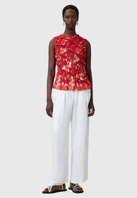 Blusa rossa texture con ruches asimmetriche, pieghe orizzontali e accenti floreali, abbinata a pantaloni bianchi larghi e sandali metallici.