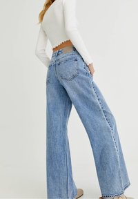 Jeans anchos de color azul claro con una cintura alta, que cuentan con bolsillos traseros y una sutil textura de lavado piedra. Estilizados con una blusa blanca de manga larga.