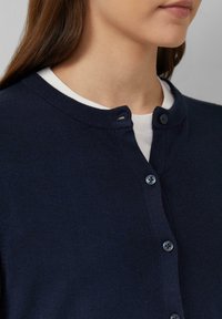 Marineblauwe cardigan met een ronde hals, slim-fit ontwerp en een knoopsluiting met zes donkere knopen. Zachte gebreide textuur en lange mouwen.