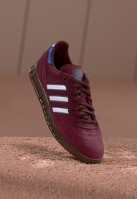 Bordeaux mockasinsneaker med blå accenter, med tre vita ränder, brun gummisula och perforerad tåhuvuddetalj.