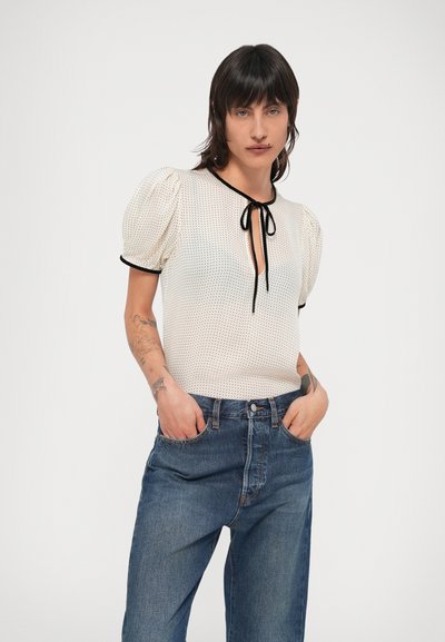 Ανοιχτό κρεμ μπλούζα με φουσκωτά μανίκια, μαύρη διακόσμηση και κορδόνι στο λαιμό, συνδυασμένη με σκούρο μπλε τζιν ψηλόμεσο παντελόνι.