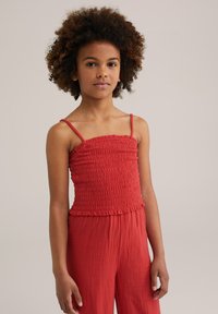Red smocked top with thin straps and ruffled hem, made of soft fabric. Model has curly hair and is facing the camera, with hands by sides.