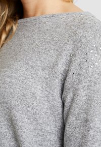 Gros plan sur un pull en maille grise orné de petits clous argentés dispersés sur l'épaule et le haut de la poitrine, porté par une personne aux cheveux blonds.