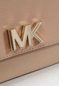 Logo MK métallique doré sur une surface en cuir beige clair avec détail de couture visible en bas du cadre.