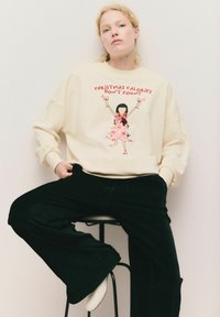 Kremowa bluza z graficzną ilustracją i napisem "Świąteczne kalorie się nie liczą". Zestawiona z czarnymi spodniami o szerokich nogawkach i białymi butami.