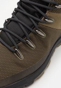 Brązowe i czarne buty trekkingowe z teksturowaną cholewą, czarnymi sznurowadłami, metalowymi oczkami i szorstką czarną podeszwą. Trwałe materiały zwiększają przyczepność.