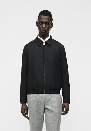 Mand iført en sort jakke med lynlås og krave samt lysegrå bukser, stående mod en ensartet lys baggrund.