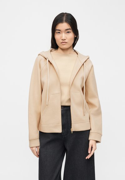 Γυναίκα με μπεζ φούτερ με φερμουάρ πάνω από μπεζ μπλούζα και σκούρο παντελόνι ψηλής μέσης, στέκεται μπροστά σε απλό λευκό φόντο.