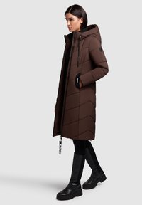 Pardoseală lungă maro cu glugă și închidere cu fermoar, având cusături în formă de V, buzunare laterale și un accent cu logo pe tiv. Botine negre până la genunchi.