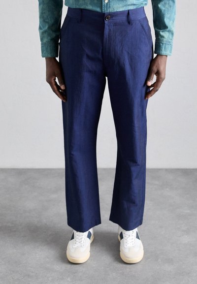 Pantalons en lin bleu marine avec une coupe droite, dotés d'une fermeture à bouton à l'avant et de poches latérales, associés à des baskets blanches et beiges.