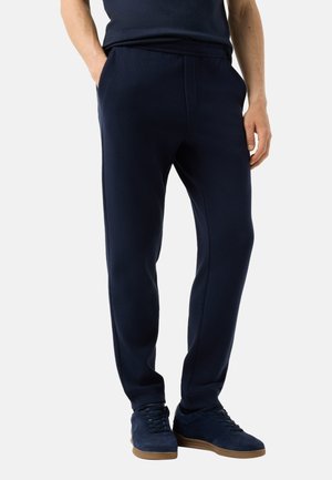 Man draagt donkerblauwe joggingbroek en bijpassende sneakers, staat met handen in de zakken tegen een effen achtergrond.