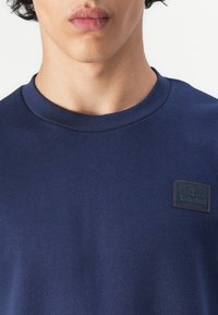 Sötétkék pulóver bordás nyakkal, fekete Timberland logó folttal a mellkasán, sima, pamutszerű anyagból készült.