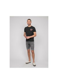 T-shirt en coton noir avec un design graphique, shorts en denim gris et chaussures légères à enfiler, posant devant un fond neutre.