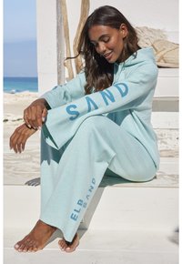Hellblaues Lounge-Set aus weichem Stoff, bestehend aus einem Kapuzenoberteil und weiten Hosen. "SAND"-Logo auf den Ärmeln und Hosen.