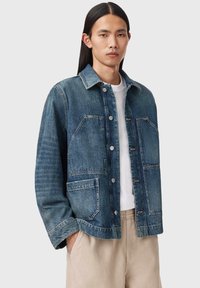 Kurtka dżinsowa w średnim odcieniu niebieskiego z przednim zapięciem na guziki, dwoma kieszeniami na piersi i długimi rękawami. Posiada klasyczny kołnierz i lekko przetartą teksturę.