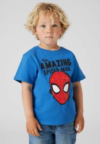 Modrá bavlnená tričko s veľkým červeným grafikom Spider-Mana, bielymi očami a čiernym textom, v kombinácii s denimovými šortkami.