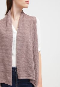 Gestrickte mauve Weste mit einem strukturierten Muster, offenem Vorderteil und ohne Verschluss. Kombiniert mit einer weißen Bluse, präsentiert vor einem neutralen Hintergrund.