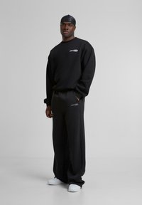 Black sweatshirt and matching wide-leg pants in fleece material. Features a small graphic logo on the chest. Paired with white sneakers.