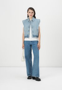 Colete de ganga com colarinho e botões frontais, combinado com uma t-shirt branca e jeans azul claros à boca de sino. Ela segura uma pequena bolsa branca, vestindo botas pretas.